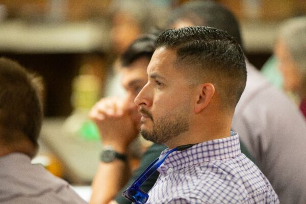 A man with short dark hair and a trimmed beard, wearing a checkered shirt and glasses around his neck, sits attentively among a blurred group of people in a meeting or conference setting.
