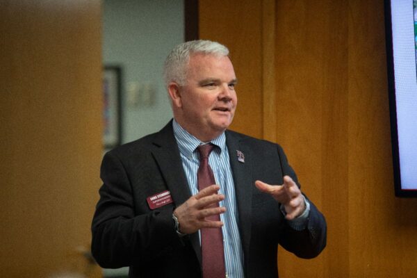A man in a dark suit and striped shirt gestures while speaking indoors. He wears a maroon tie and a name tag, standing in front of a wooden wall and a partially visible screen.