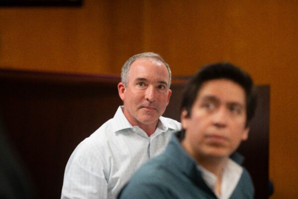 Two men indoors, both wearing collared shirts. The man in the background is looking forward with a neutral expression, while the man in the foreground is slightly out of focus and looking to the side.