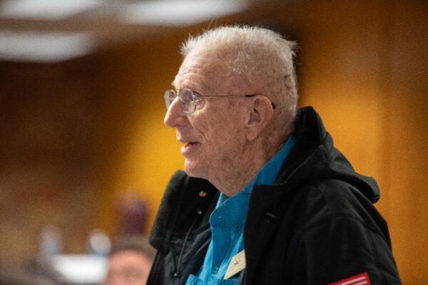 An elderly man with short white hair, glasses, and a teal shirt under a black jacket stands indoors, smiling and looking to the left. The background is softly blurred with warm lighting.