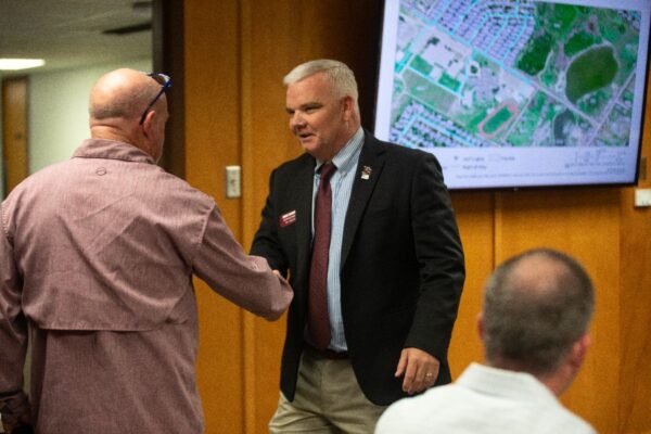 A man in a suit and tie shakes hands with another man wearing a casual shirt, while a third person sits in the foreground. Behind them is a large screen displaying a map with streets and green areas.