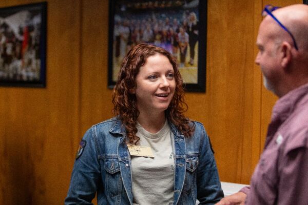 A woman with curly red hair wearing a denim jacket and name tag talks to a bald man in glasses and a purple shirt in a room with wooden walls and framed photos.