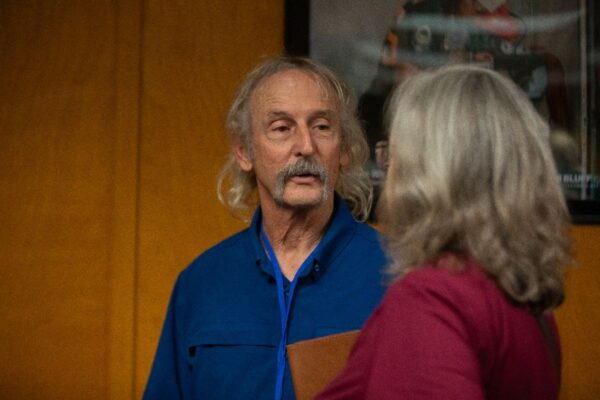 A man with long gray hair and a mustache, wearing a blue shirt and holding a folder, talks to a woman with gray hair in a red top. They are indoors with a yellow wall and framed picture in the background.