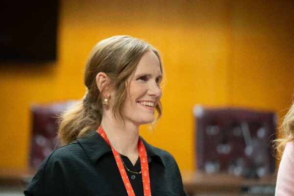 A woman with light brown hair in a ponytail, wearing a black shirt and red lanyard, smiles in an indoor setting with a blurred orange and brown background.