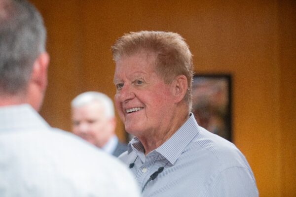 A smiling older man with short, light hair and a striped shirt stands indoors, facing another person. The background is softly blurred with two other people visible.