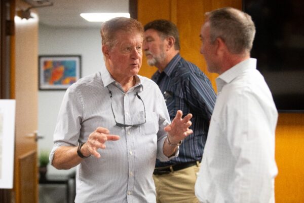 An older man gestures while talking to another man in an office setting, with a third man standing in the background. All are dressed in business casual attire.