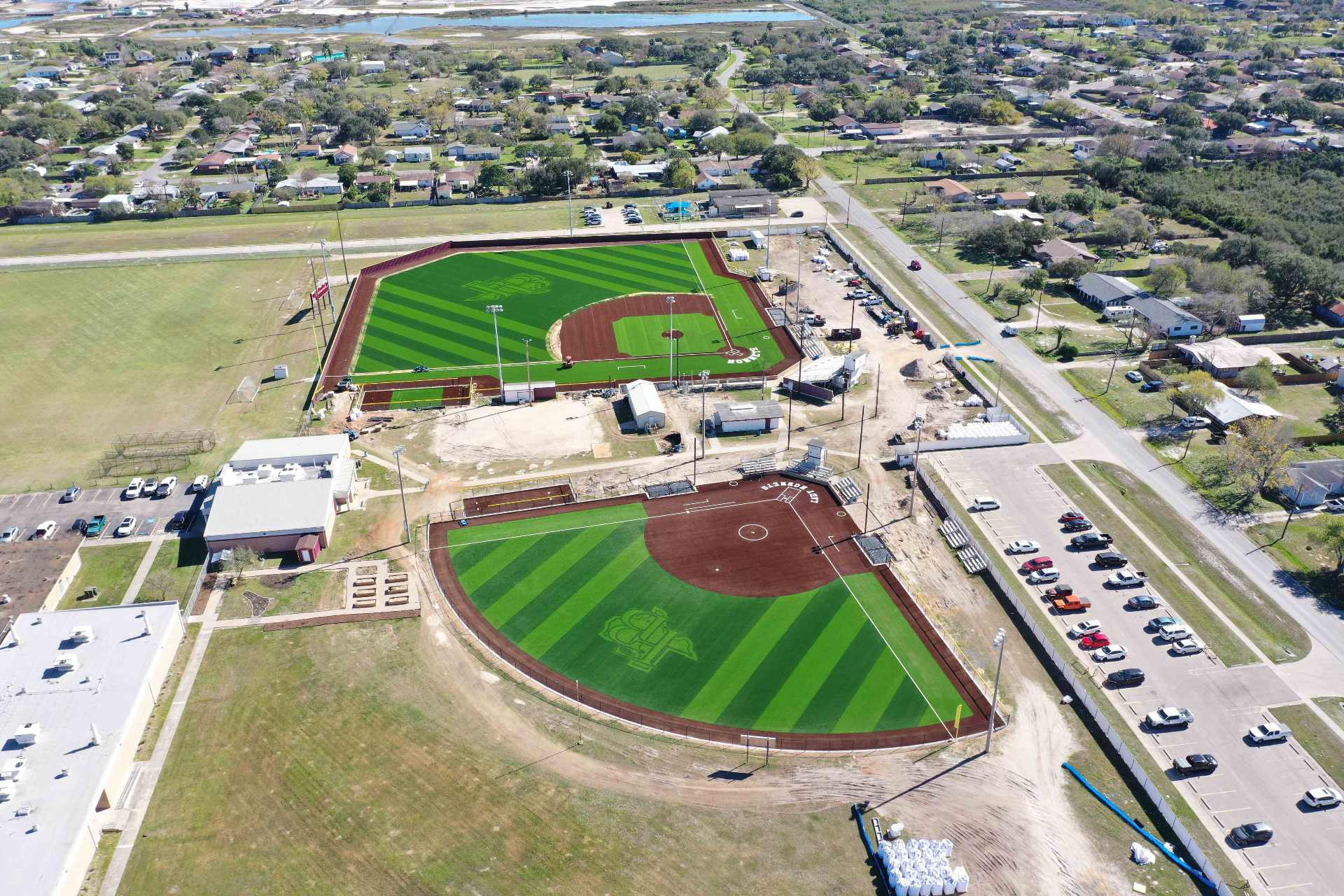 Aerial view of two adjacent baseball fields with green turf and brown infields, parking lots with cars, surrounding roads, nearby buildings, and a residential neighborhood in the background.