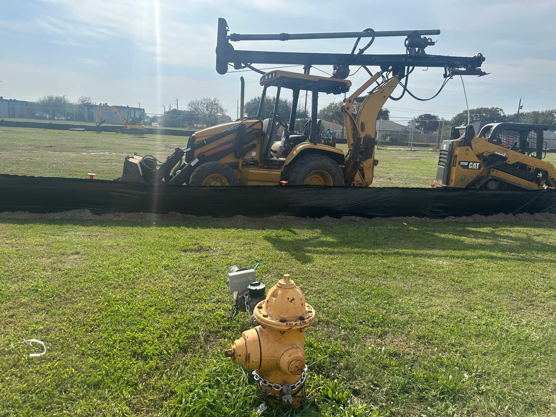 A yellow fire hydrant chained to the ground is in the foreground, with construction equipment, including a backhoe and a skid steer, behind a black silt fence on a grassy lot under a partly cloudy sky.
