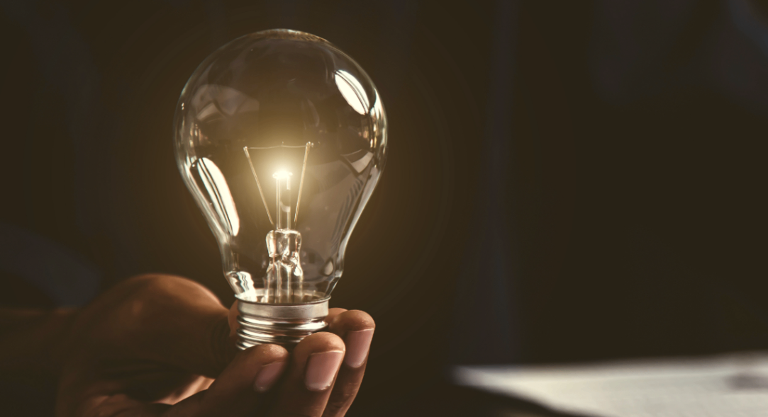 A person’s hand holds a glowing light bulb against a dark background, symbolizing a bright idea or creativity. The bulb is gently gripped, and light softly illuminates the surrounding area.