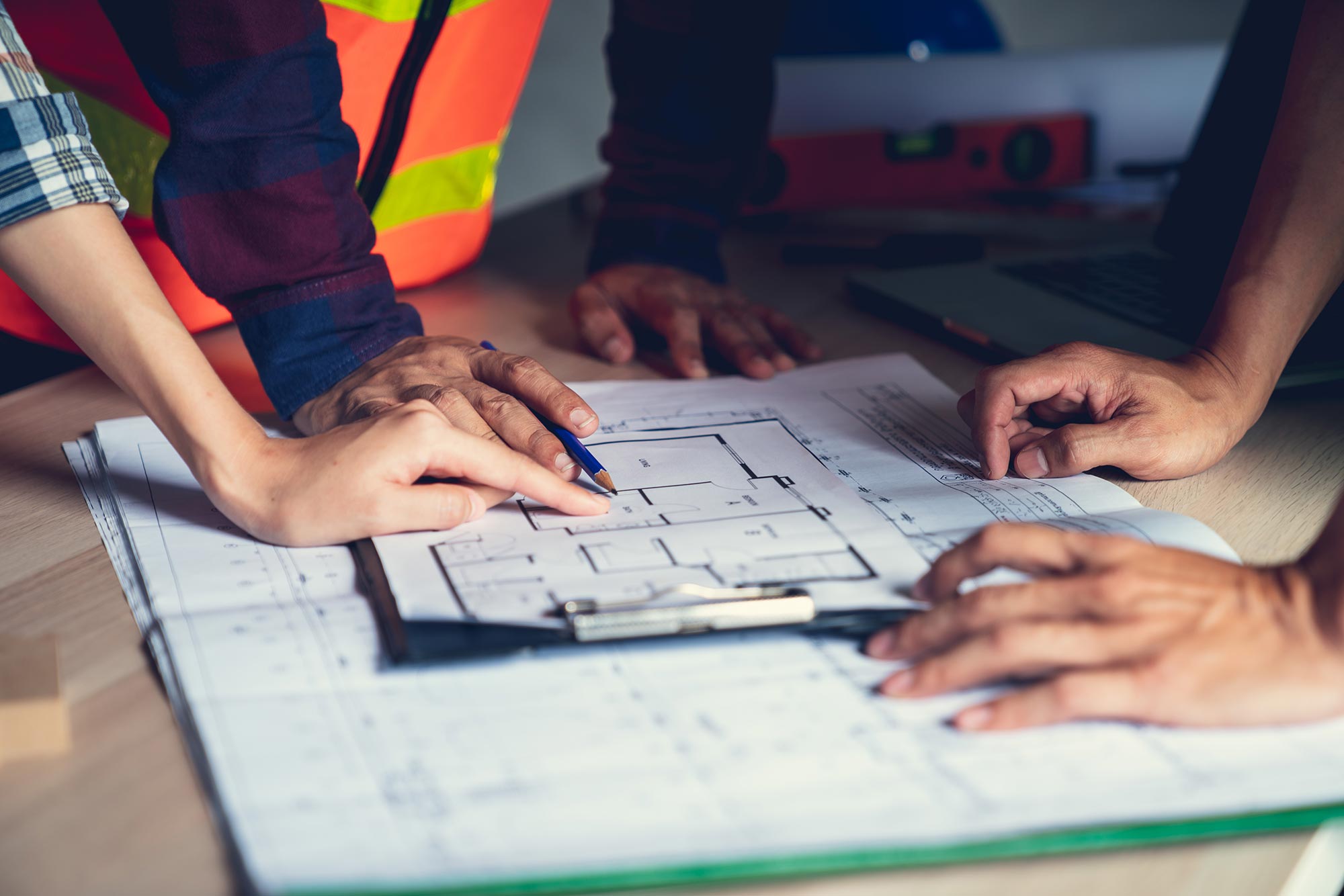 Three people review architectural blueprints spread on a table. One points at the plans with a pen, while another’s hand rests on the paper. A clipboard and laptop are visible; one person wears a hi-vis vest.