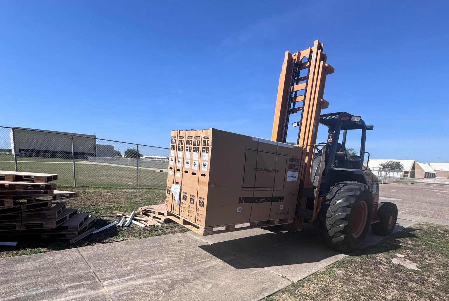 A forklift lifts and transports large cardboard boxes on a paved path near a fenced grassy area, with buildings visible in the background under a clear blue sky.