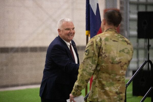 A man in a dark suit and tie shakes hands with a uniformed soldier in camouflage, standing indoors near a flag and a small stage. The man in the suit is smiling warmly.