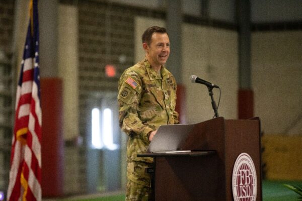 A person in military uniform stands at a podium with a microphone, smiling. The podium has an emblem, and an American flag is visible in the background inside a large indoor setting.