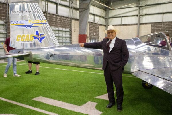 A man in a suit and cowboy hat smiles while standing indoors beside a shiny, silver small aircraft labeled Tango Flight and CCAD. The scene is on artificial turf inside a large building.