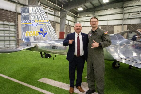 Two men stand smiling and posing together in front of a small silver airplane inside a hangar; one is in a suit and the other in a flight suit. The plane has Tango Flight and CCAD logos on its tail.