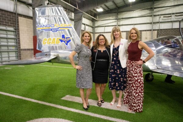 Four women stand smiling in front of a small silver aircraft inside a hangar with artificial turf. The plane’s tail displays Tango Flight, CCAD, and Splendid Aviation logos.