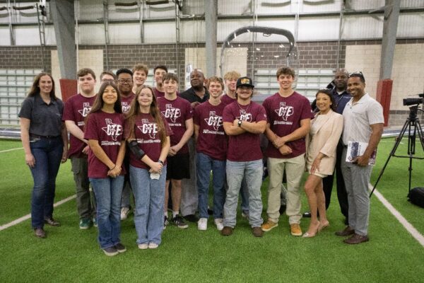 A group of students in maroon shirts with GTE logos and several adults stand together on indoor turf, smiling at the camera. Some adults hold papers; a camera on a tripod is visible in the background.