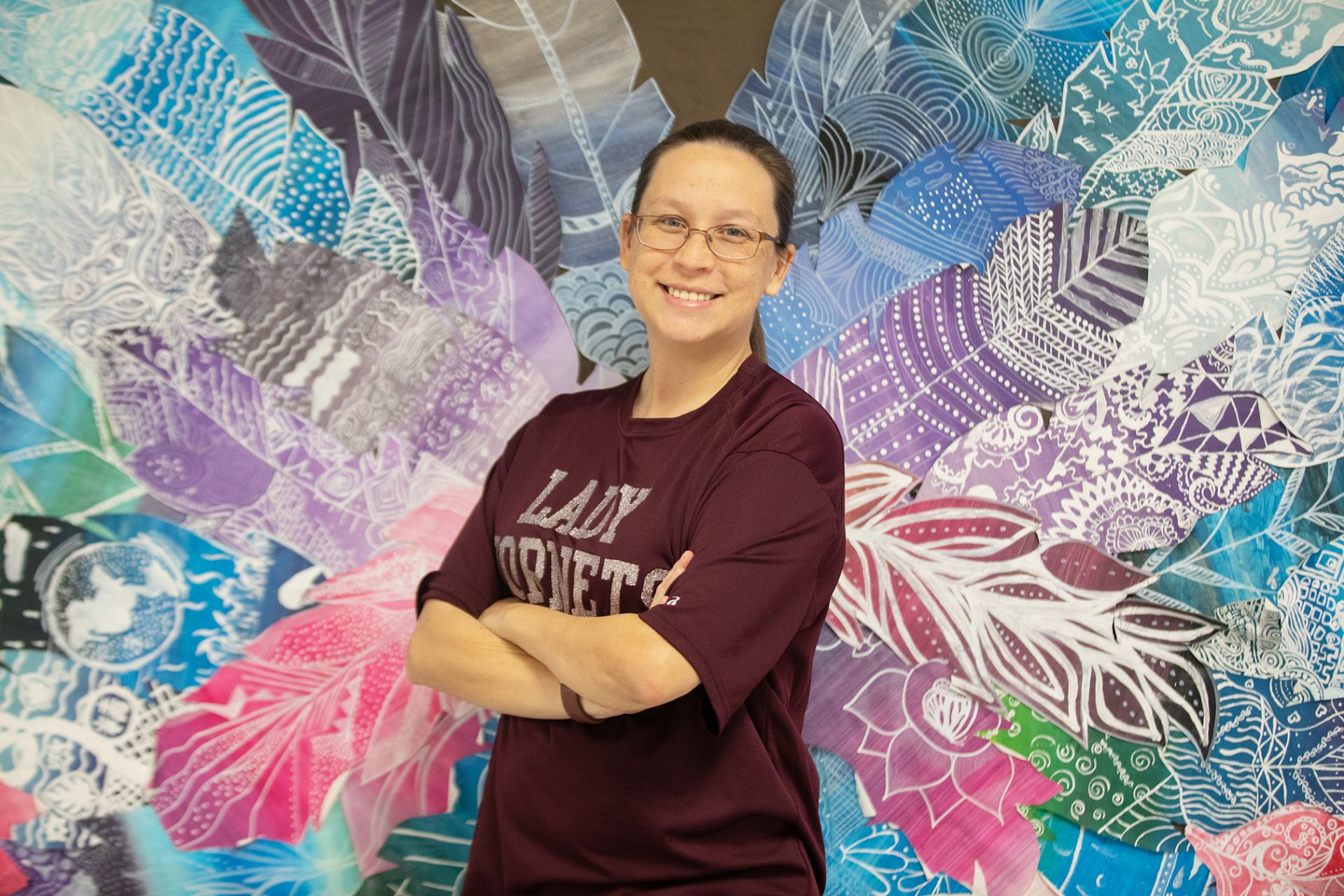 Audreana Underbrink - Principles of Art, Audio & Video Technology A smiling woman in glasses and a maroon Lady Hornets shirt stands with arms crossed in front of a vibrant, colorful mural featuring layered feather and leaf patterns in blue, purple, and pink shades.