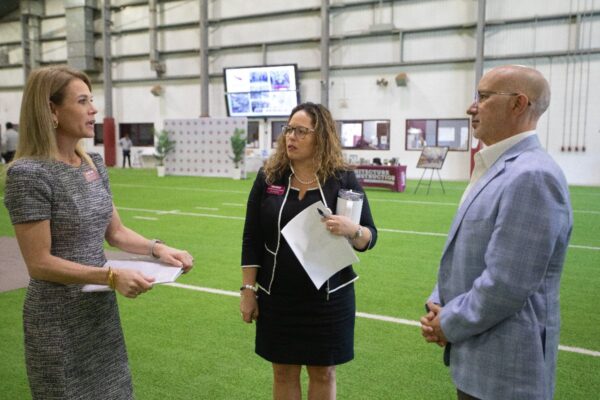 Three people in business attire talk while standing on an indoor sports field. Two women hold papers, and a man in a light suit listens. Behind them are sports equipment, a photo display, and a large screen.