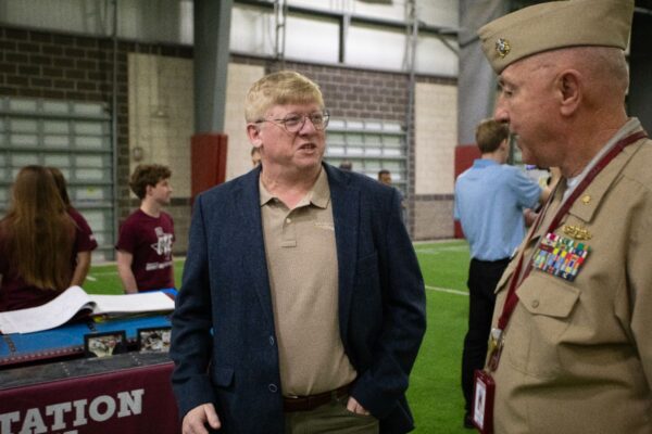 A man in a blue blazer speaks with a uniformed military officer inside an indoor facility. People and a display table with photos and open books are visible in the background.