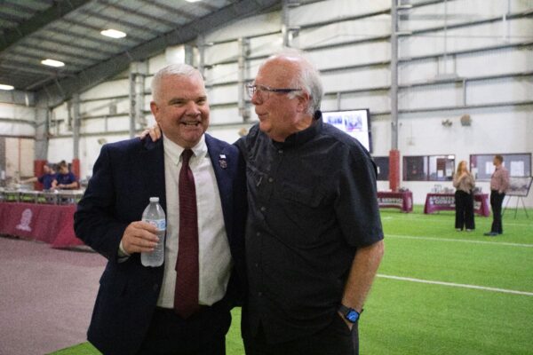 Two men stand indoors on artificial turf, smiling and talking. One wears a suit and holds a water bottle, while the other wears a dark shirt and glasses. Tables and people are visible in the background.