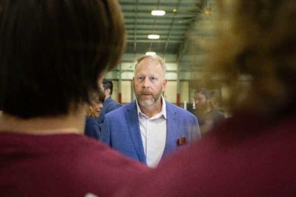 A man in a blue blazer stands indoors, facing two people with their backs to the camera, engaging in conversation. The setting appears to be a large, well-lit hall or warehouse.