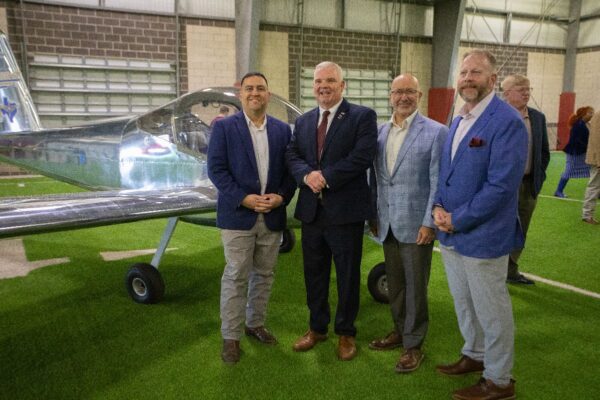 Four men in suits stand smiling in front of a small silver airplane inside a large indoor space with artificial turf and industrial walls. Other people are visible in the background.