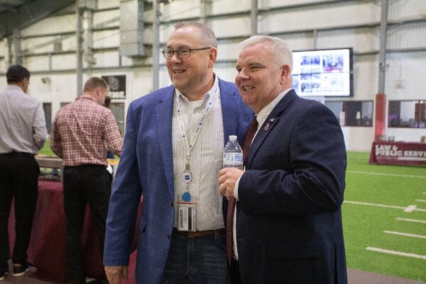 Two men in business attire smile and pose together at an indoor event on artificial turf. One holds a water bottle, while people and tables are visible in the background. Large screens display images on the wall behind them.