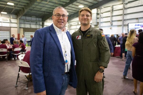 Two men pose and smile for a photo indoors at an event. One wears a blue blazer and glasses, the other is in an olive green military flight suit. People are seated and mingling in the background.