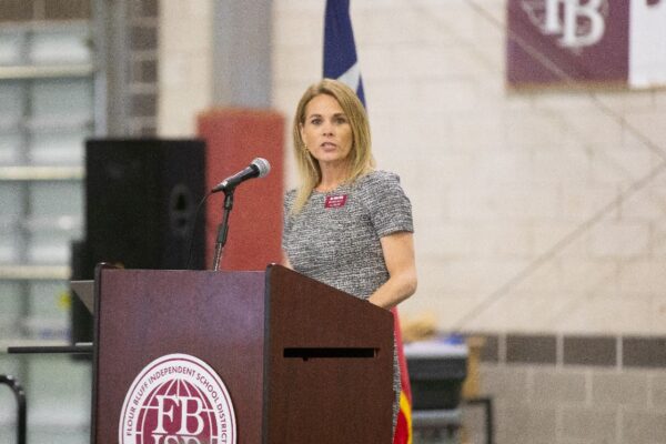 A woman stands at a podium, speaking into a microphone in a gymnasium. She wears a gray dress, and a badge. A school district logo and banners are visible in the background.