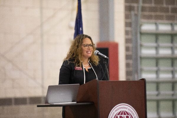 A woman with curly hair, wearing glasses and a black blazer, speaks at a podium with a laptop on it. There is a microphone and an American flag in the background. The podium displays a school district logo.
