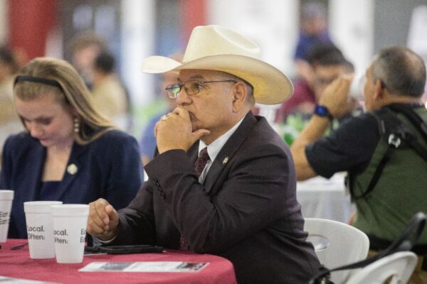 A man in a cowboy hat and suit sits at a round table with white cups and papers, thoughtfully resting his chin on his hand. People are seated around him at what appears to be an indoor event or conference.
