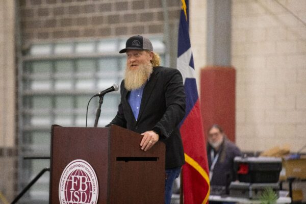 A bearded man in a black jacket and cap speaks at a podium with the FBISD logo. An American flag and another person sitting at a table are visible in the background.