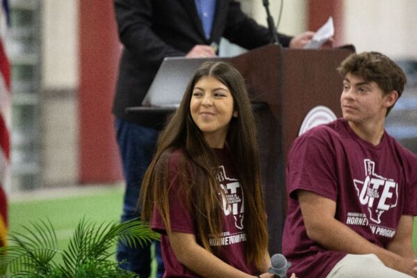 Two students in matching maroon T-shirts sit and smile at an indoor event, with a person speaking at a podium in the background and an American flag partially visible to the left.