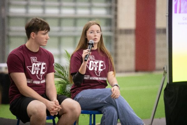Two young people wearing matching maroon CTE Texas shirts sit on stools indoors. The woman holds a microphone, speaking, while the man listens. They are beside a plant and a projector screen, with a blurred background.