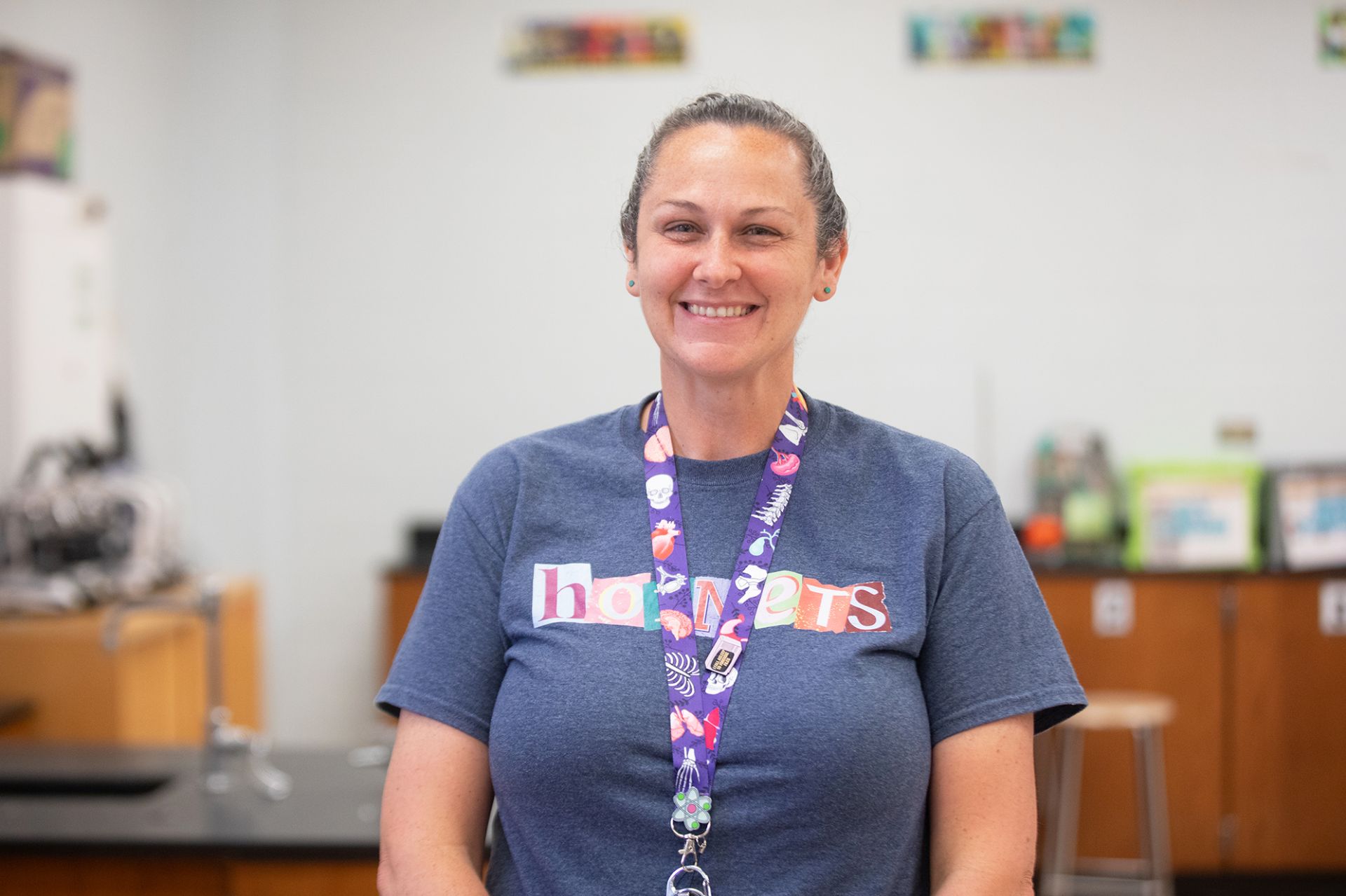 Brandi Venable - Forensic Science A woman with brown hair tied back, wearing a blue hornets t-shirt and a colorful lanyard, smiles while standing in a classroom with science equipment in the background.