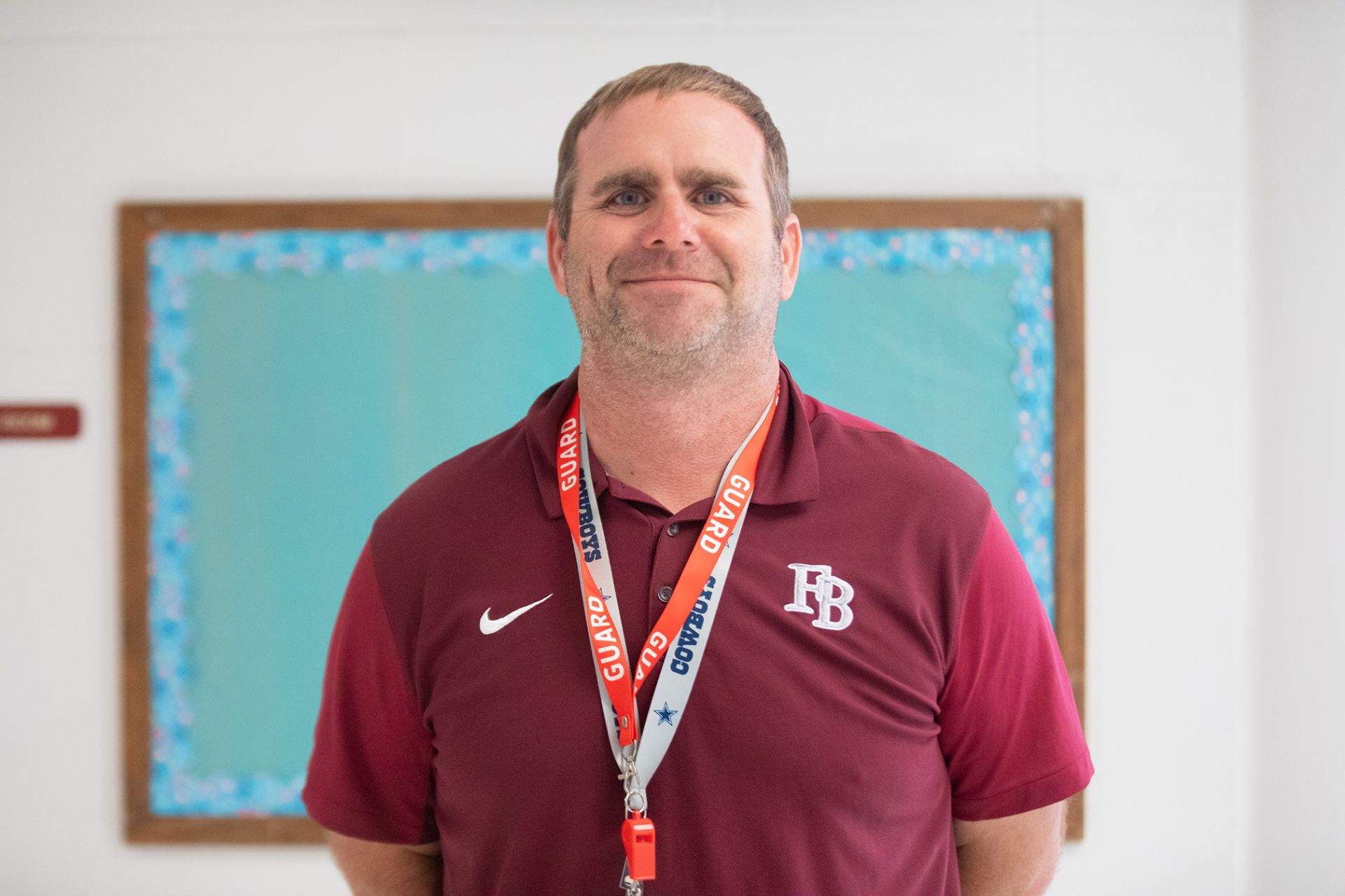 Brett Boyd - Principles of IT A man wearing a maroon polo shirt with FB initials stands indoors, smiling. He has a whistle and a lanyard around his neck, and a bulletin board with a blue background is behind him.
