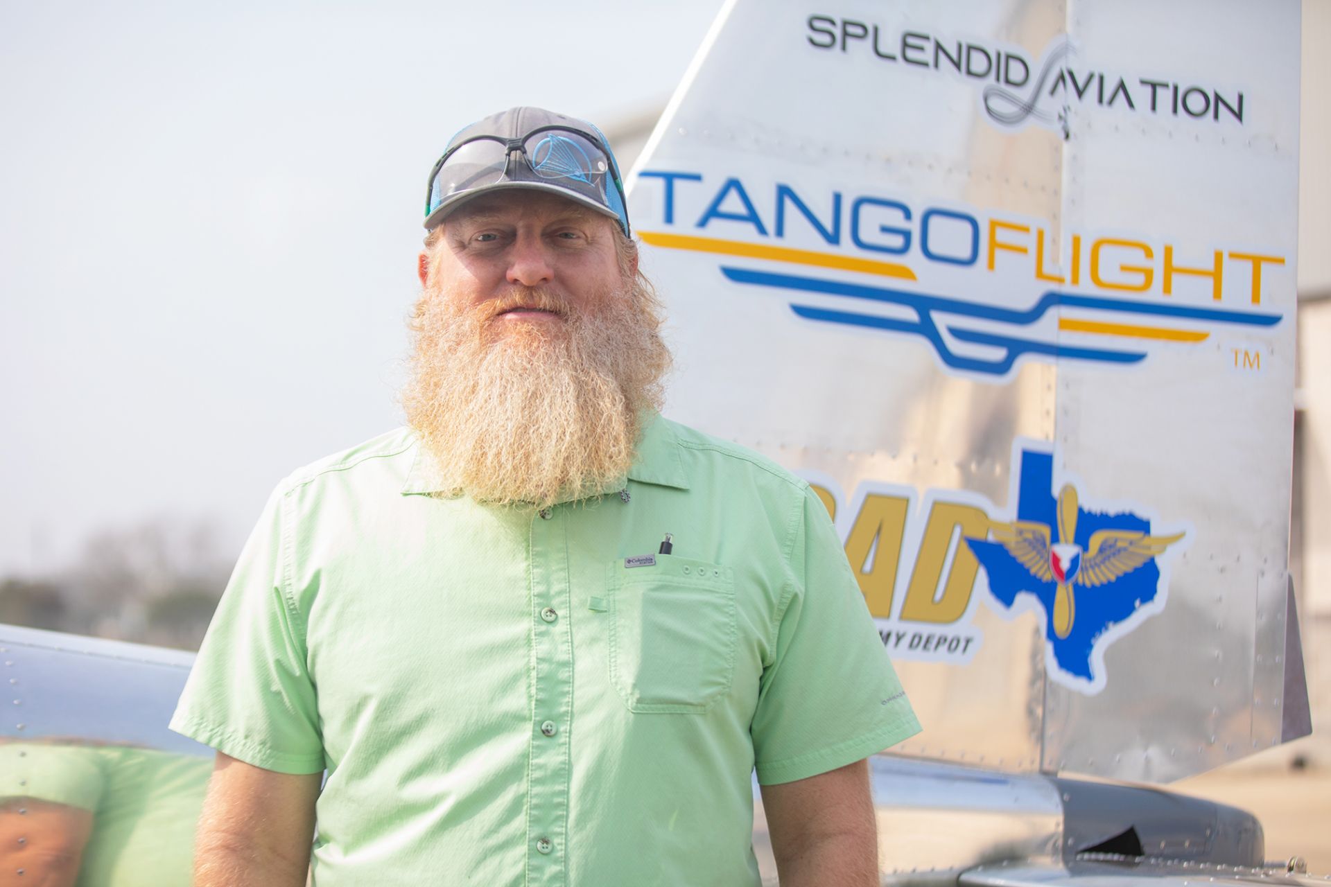 Caleb Harris - Hornet Aviation A man with a long beard, wearing a light green shirt, sunglasses, and a cap, stands in front of a shiny aircraft tail with Tango Flight and aviation logos visible.