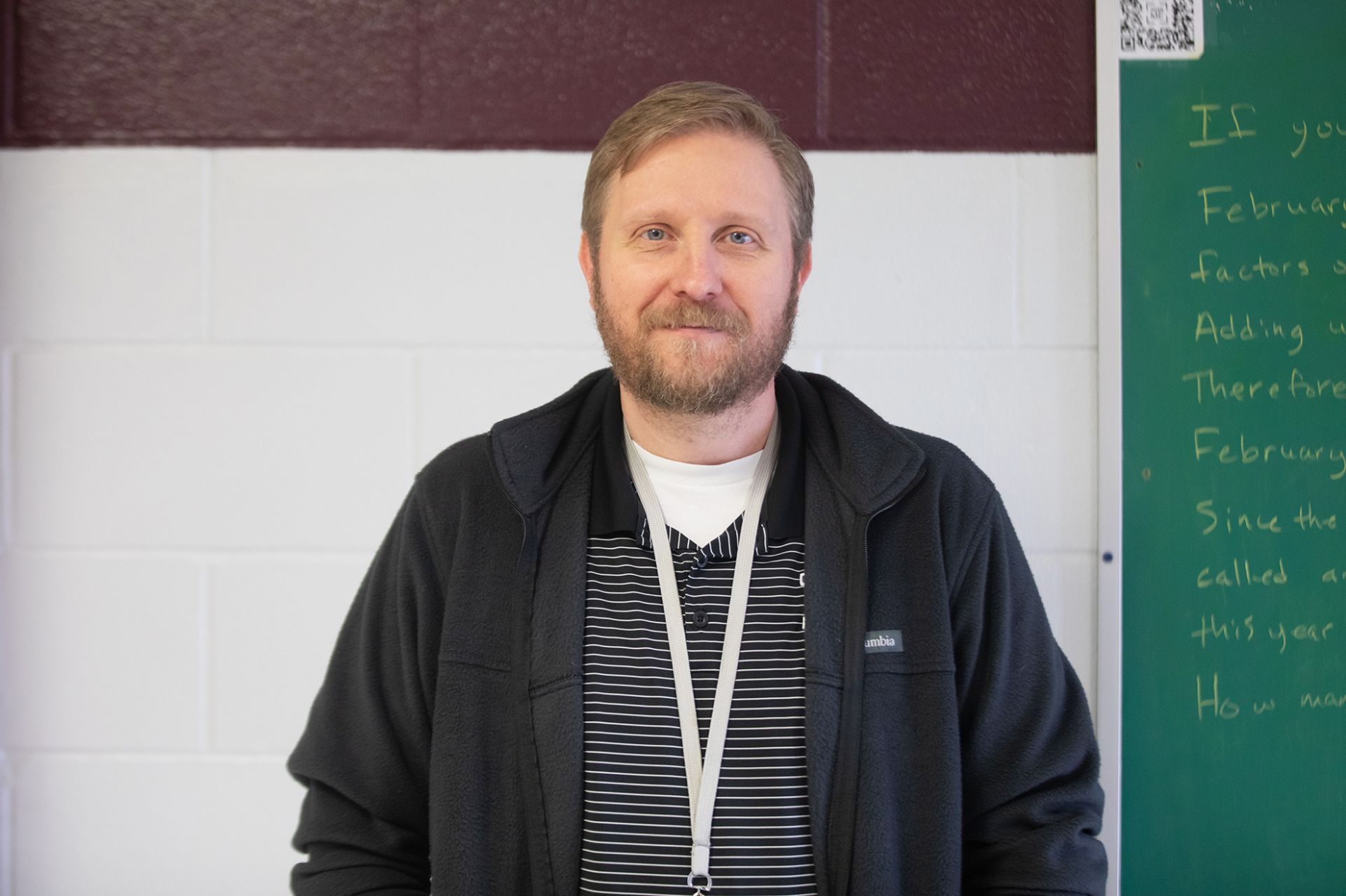 Chad Purdy - Computer Science A man with light brown hair and a beard, wearing a black jacket and striped shirt, stands in front of a white and maroon wall with part of a green chalkboard visible on the right.
