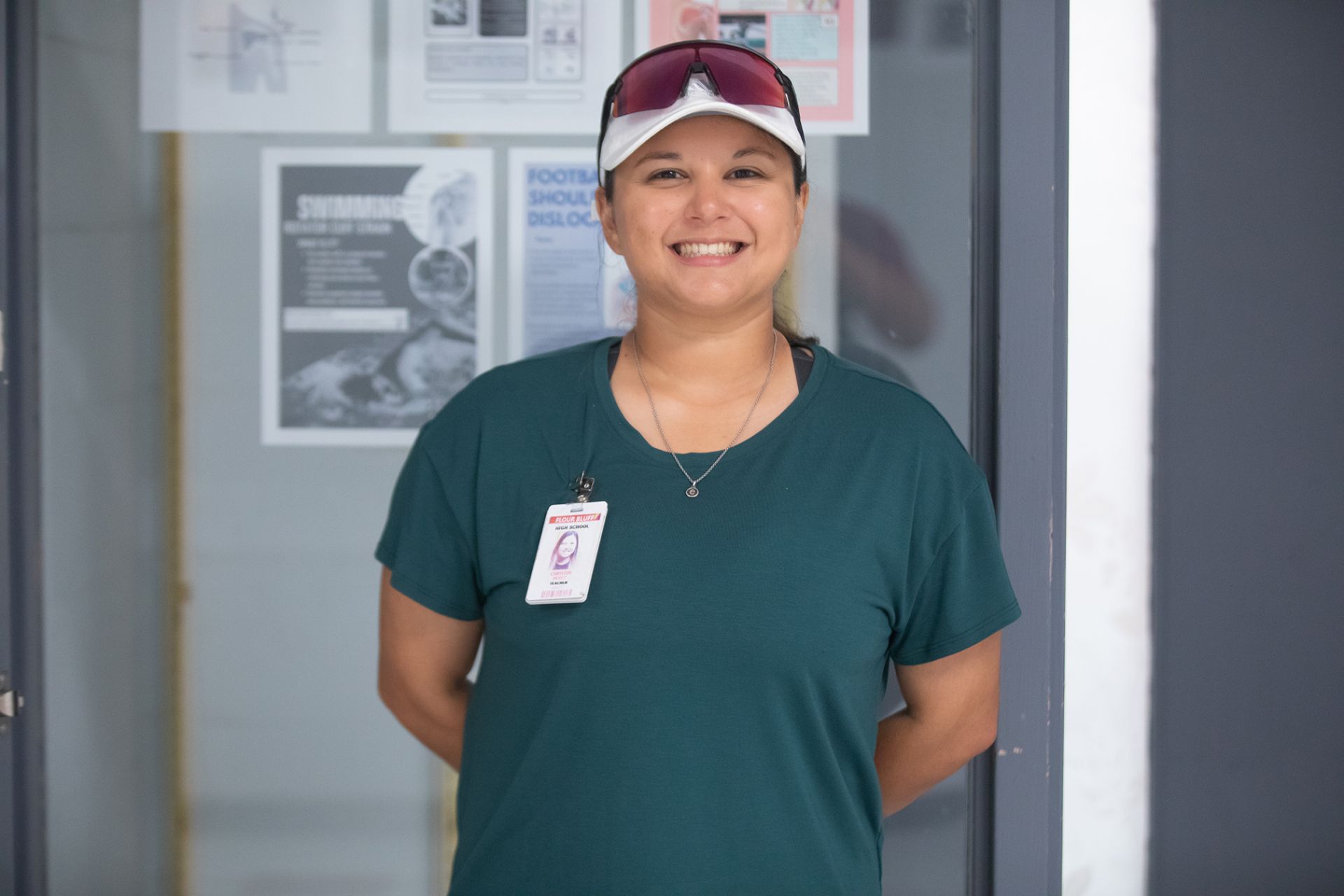 Christen Perez - Nutrition & Exercise Science A smiling person wearing a green shirt, white cap, sunglasses, and a lanyard with an ID badge stands indoors in front of a glass display case with posters.