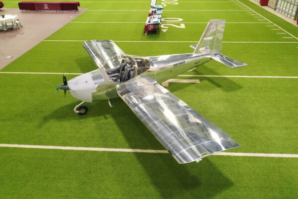 A shiny silver single-engine airplane is displayed indoors on artificial turf, near the 20-yard line of what appears to be a sports facility or stadium. Tables with maroon tablecloths are visible in the background.