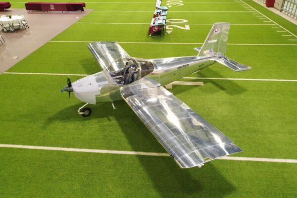 A shiny silver airplane is parked indoors on a football field with artificial turf. Tables and chairs are set up nearby along the sideline, marked with yard numbers.