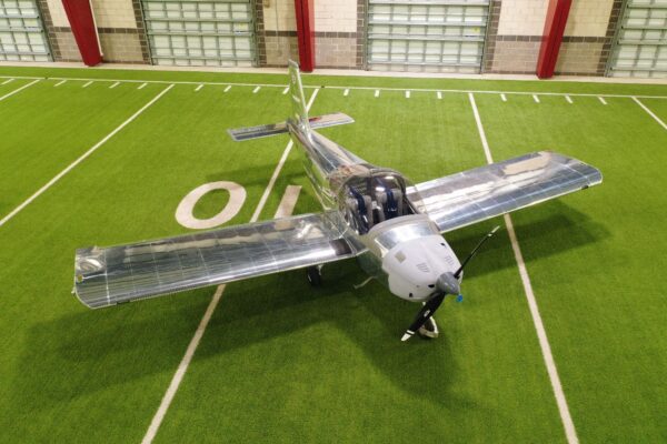 An airplane is parked indoors on artificial turf, near a white 10 marking, inside a building with large garage doors and red support beams.