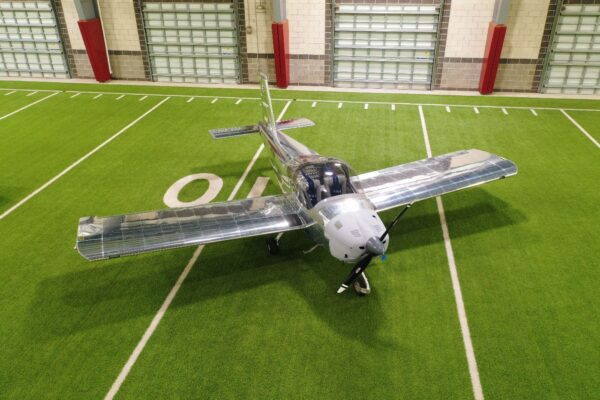 A silver aircraft with transparent wings is parked indoors on green artificial turf marked with white yard lines, resembling a football field. The planes nose is facing toward the camera.