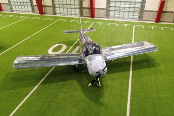 An aircraft with visible internal components is displayed on artificial turf inside a building, near a wall with large garage doors. The field markings suggest an indoor sports facility.