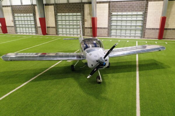 A single-engine airplane is parked indoors on an artificial turf field, with garage-style doors and red padding on the walls in the background.