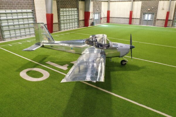 A silver aircraft is parked indoors on a green artificial turf field, near a large 40 marking. The space has high ceilings, brick walls, and several large windows and doors.