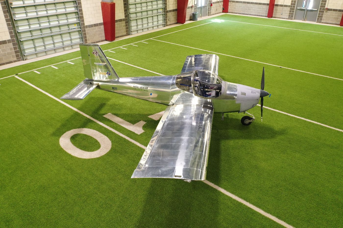 A shiny silver airplane is parked indoors on artificial turf near a white “10” yard line, inside a large building with brick walls and big windows.