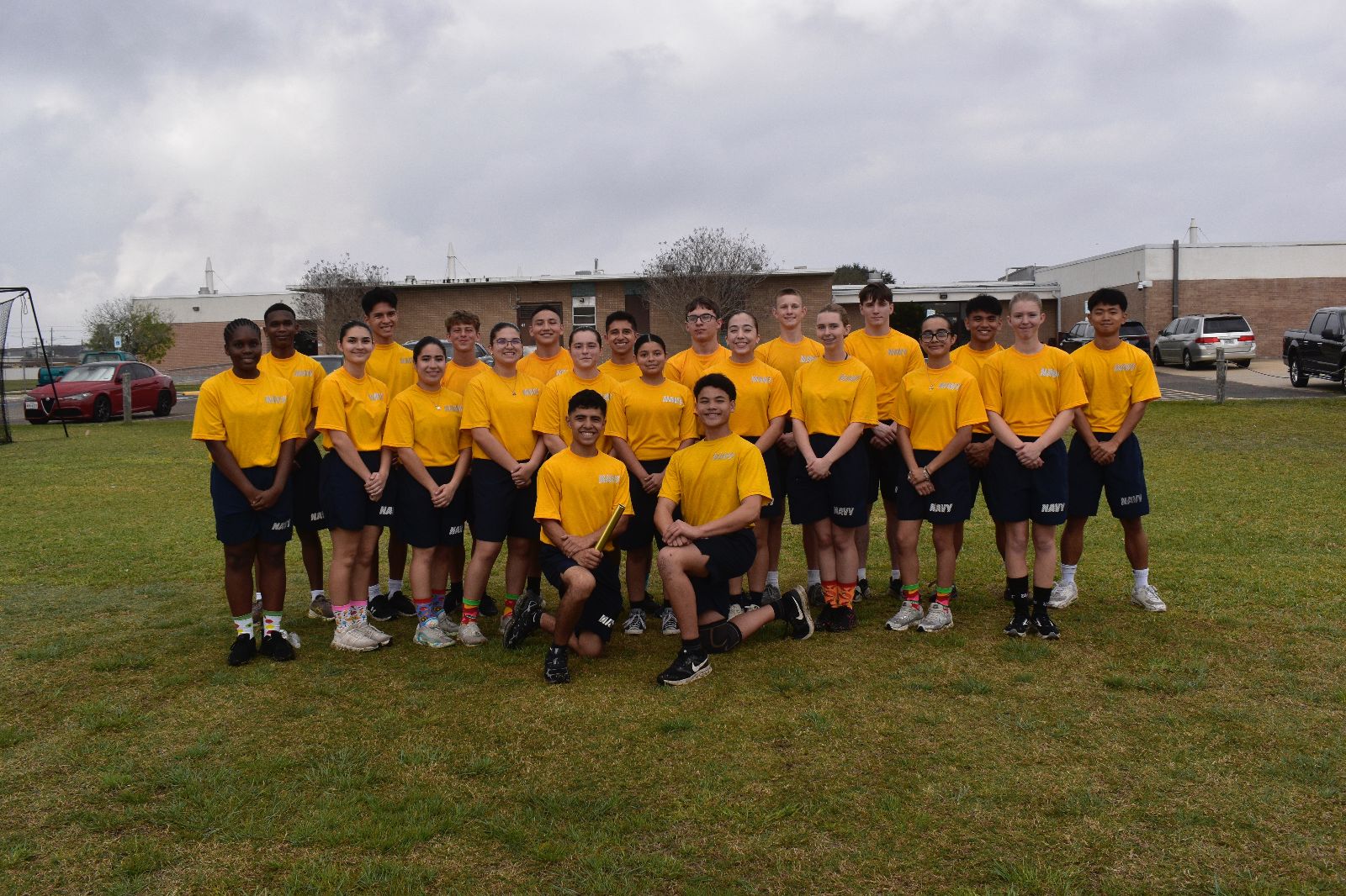 A group of young people wearing matching yellow Navy t-shirts and dark shorts pose together outside on a grassy field under a cloudy sky, with buildings and cars in the background.