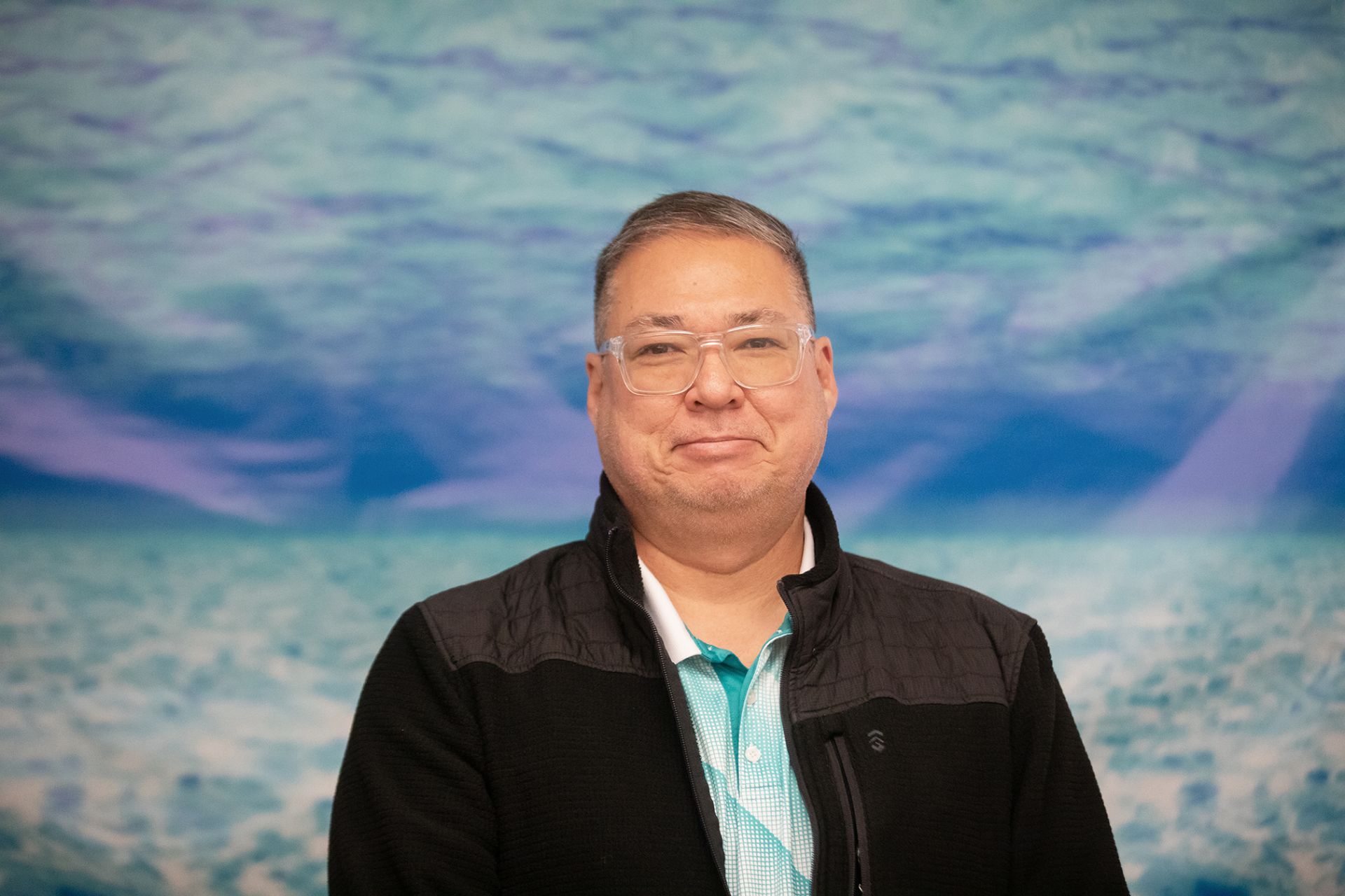 George Henderson - College & Career Exploration A man wearing glasses and a black jacket over a turquoise shirt stands smiling in front of a blue and turquoise water-themed background.